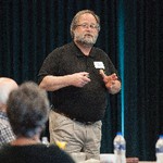 A man holding a wireless mouse gestures in front of a black curtain